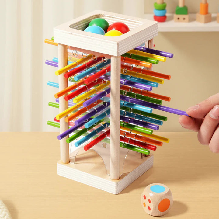 A hand pulling a purple stick from a wooden tower while colored balls rest at the top.