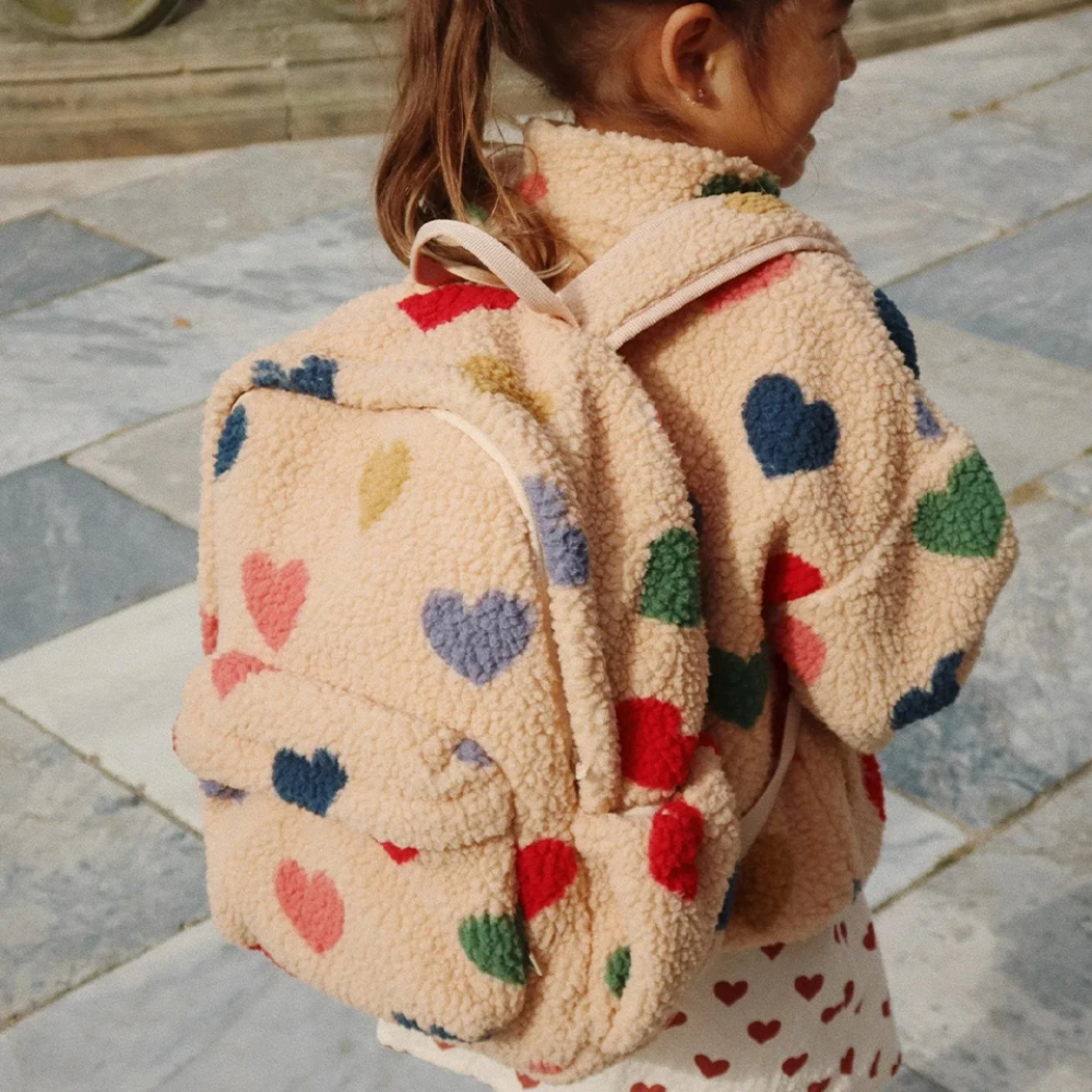 Child wearing a beige teddy backpack with multicolored hearts outdoors