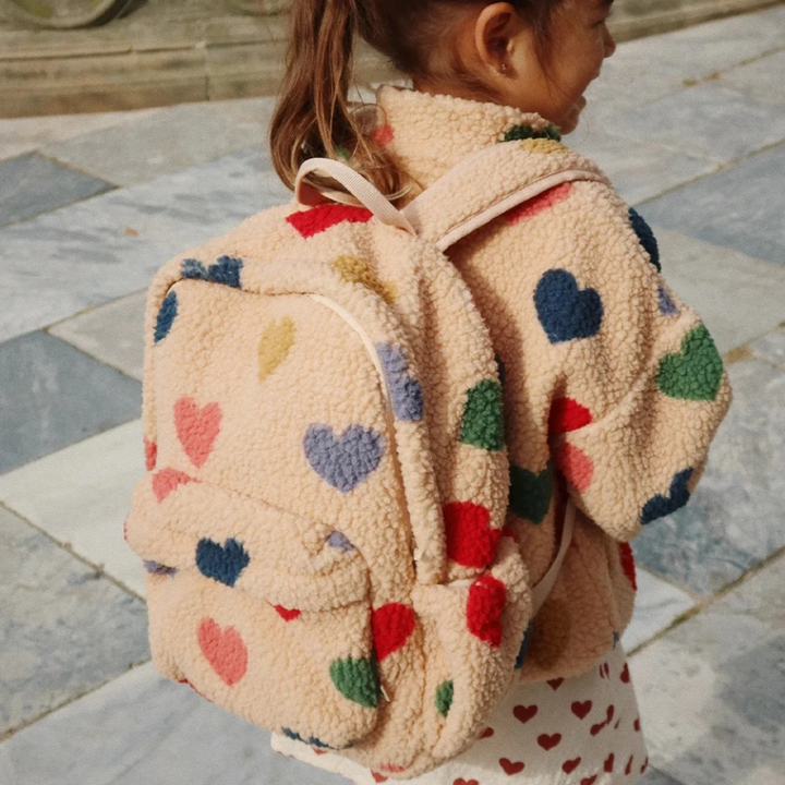 Child wearing a beige teddy backpack with multicolored hearts outdoors