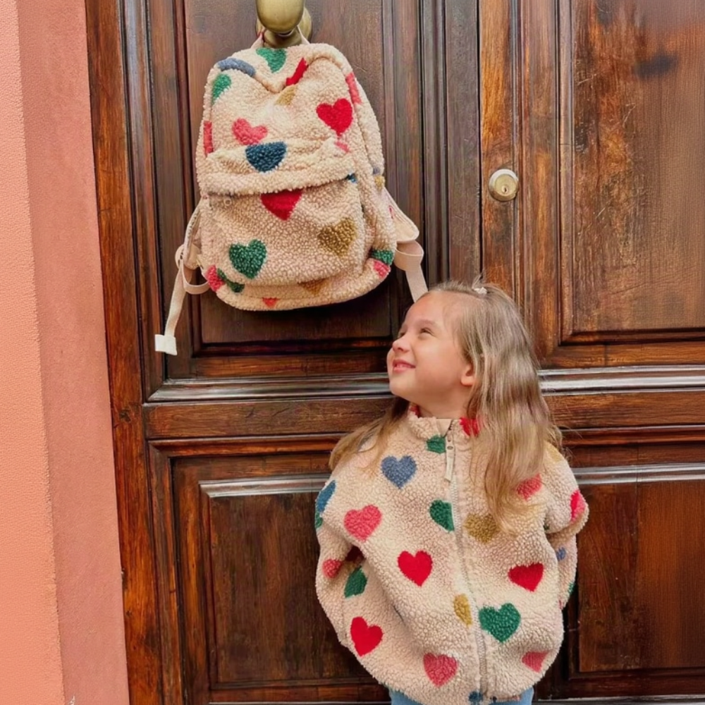 Child in a heart-patterned jacket beside a matching beige teddy backpack hanging on a door