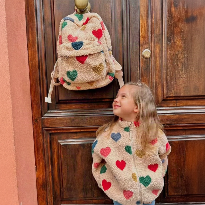 Child in a heart-patterned jacket beside a matching beige teddy backpack hanging on a door