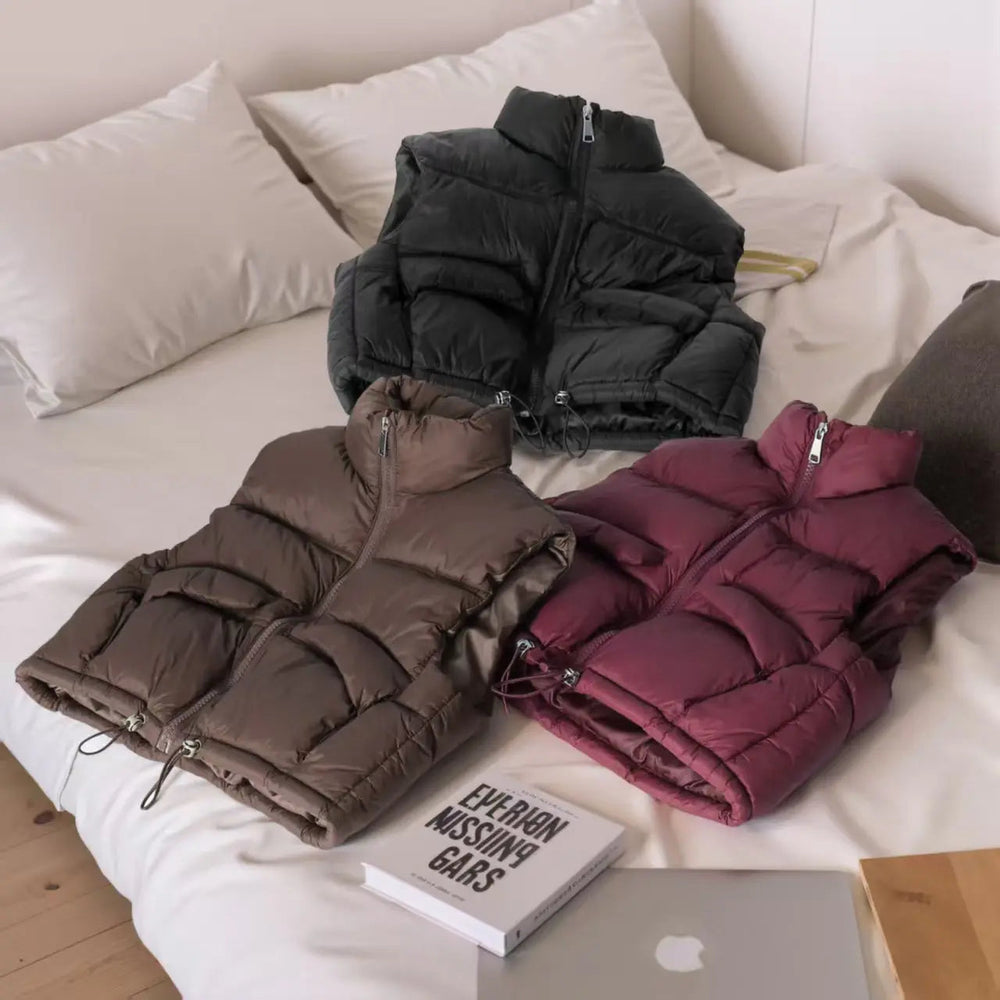 Black, Coffee, and Bordeaux Red kids' puffer vests arranged on a bed