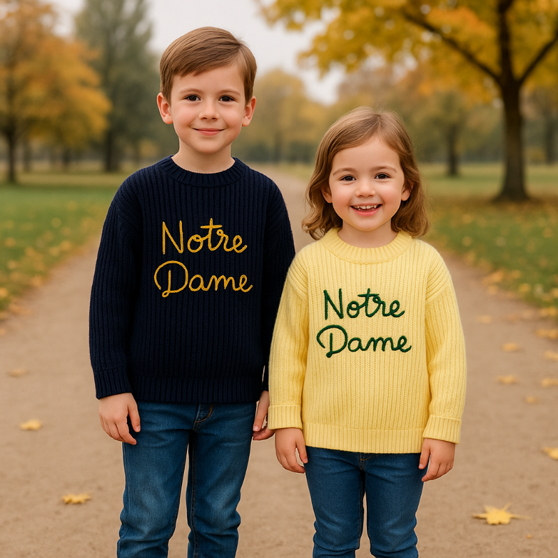 Two children wearing navy blue and yellow script-embroidered sweaters outdoors.