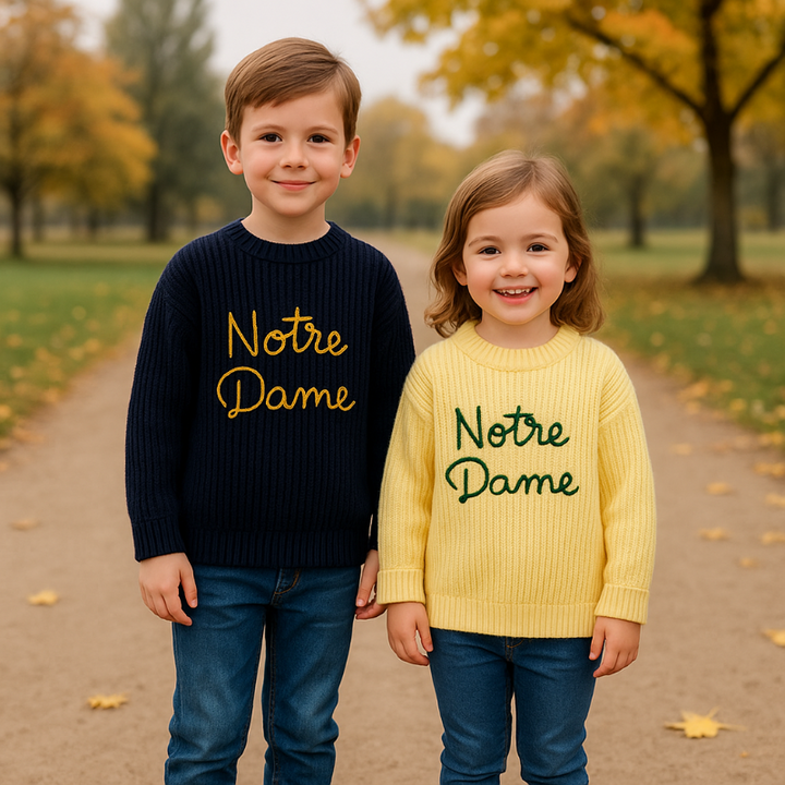Two children wearing navy blue and yellow script-embroidered sweaters outdoors.