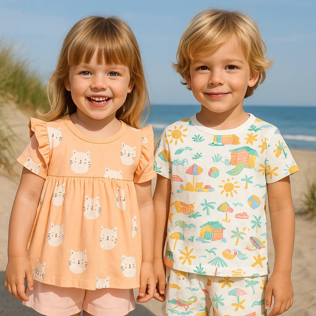 Two children wearing a peach cat-print outfit and a white seaside-print shorts set at the beach