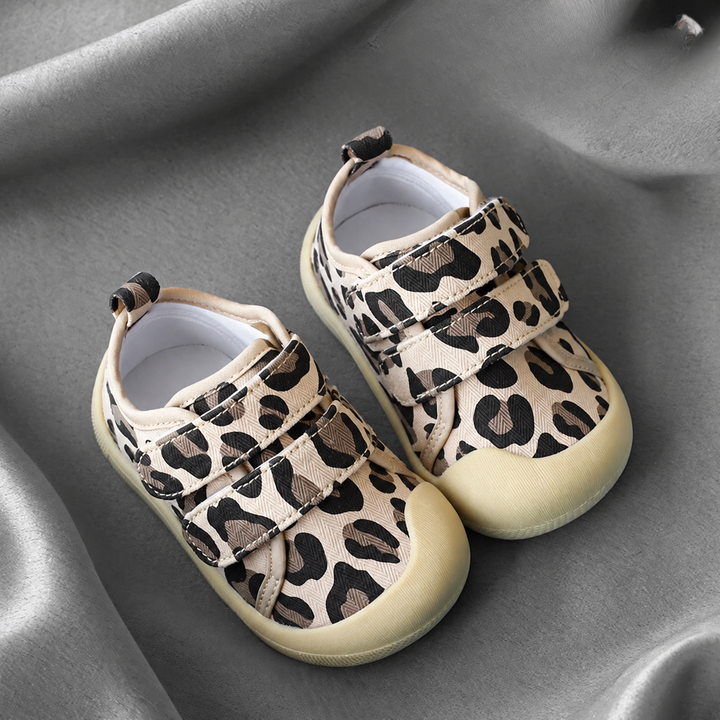Leopard-print baby sneakers with tan toe caps and strap closures on gray fabric