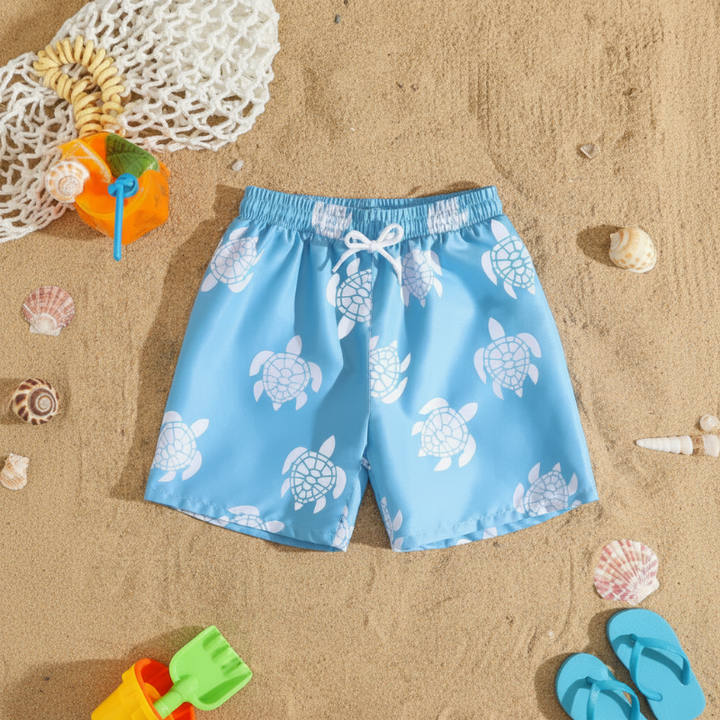 Light blue swim trunks with white turtle prints and a white drawstring laid on beach sand