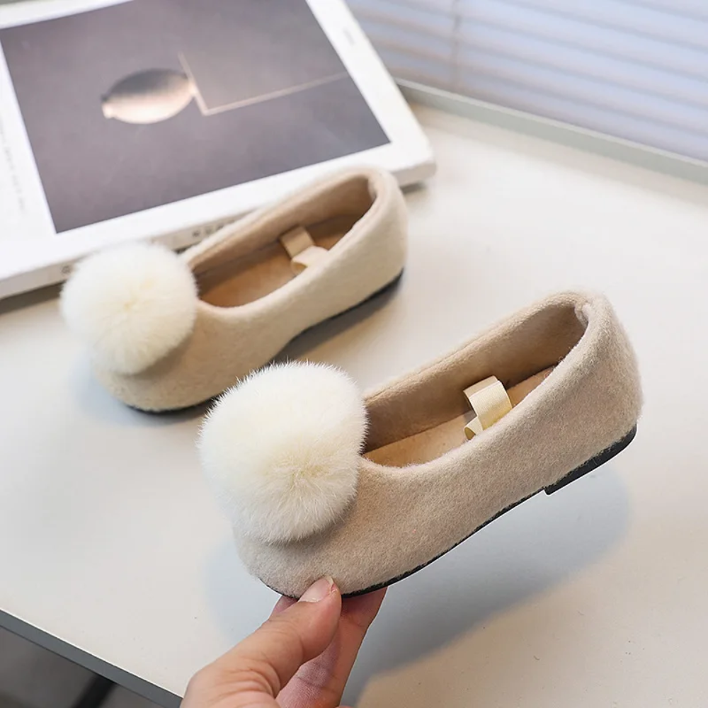 Light-colored pom-pom flats with elastic straps, with one shoe held to show the side