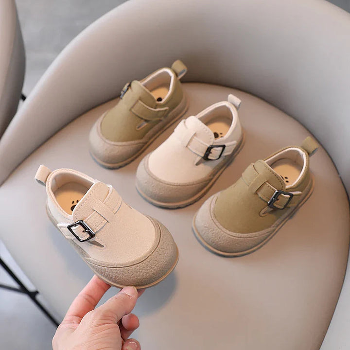 Beige baby shoe held in front of khaki and beige shoes on a chair