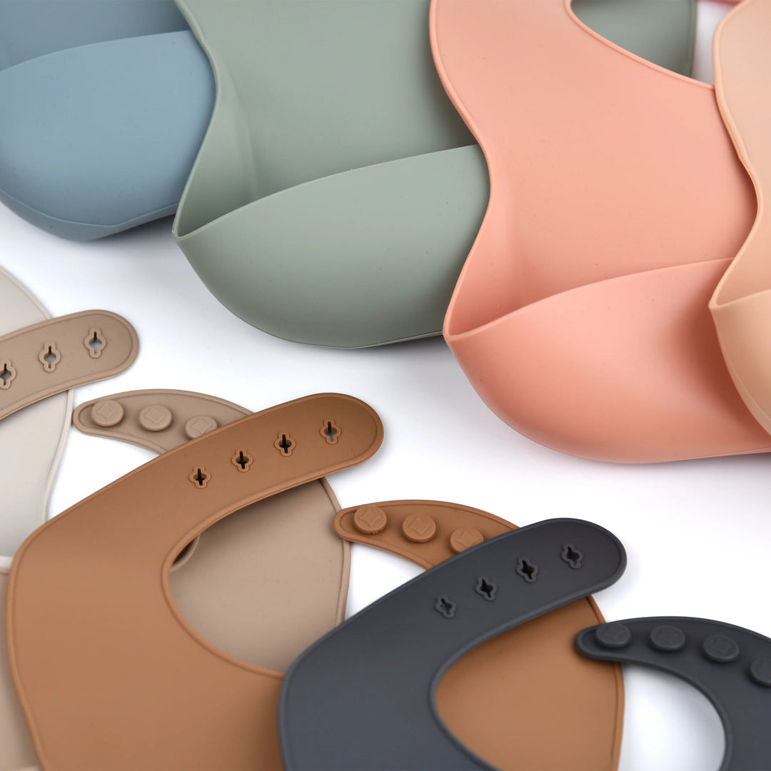 Assorted silicone bibs showing muted colors, front pockets and adjustable neck closures.