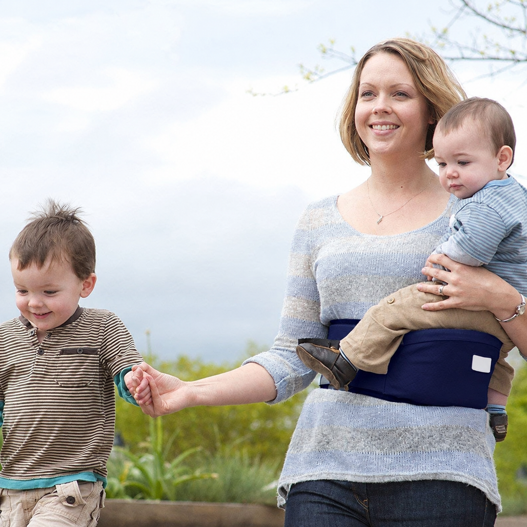Adult holding a child on a navy hip carry seat while walking with another child outdoors