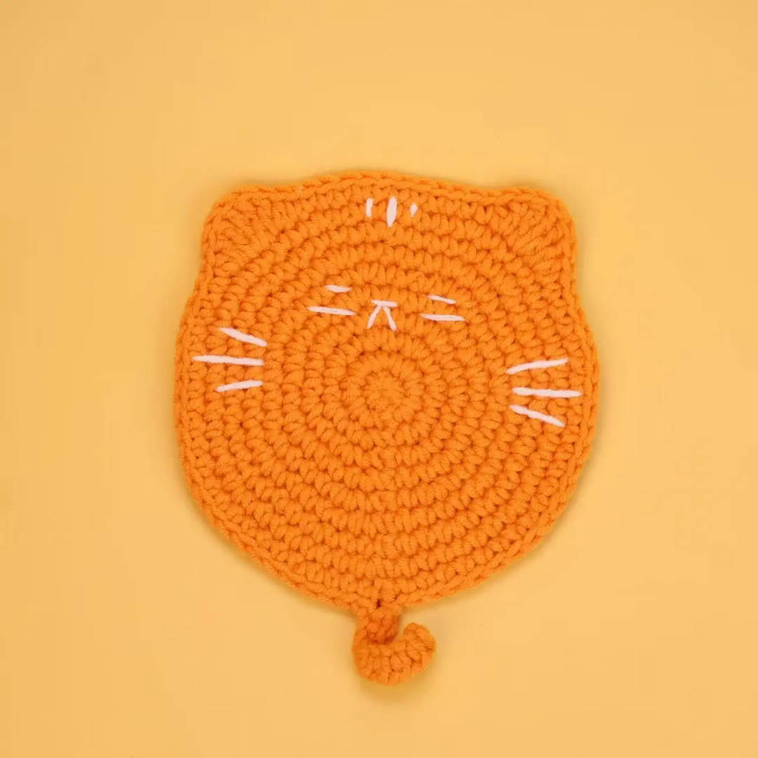 Orange crocheted cat coaster with white stitched whiskers and a curled tail