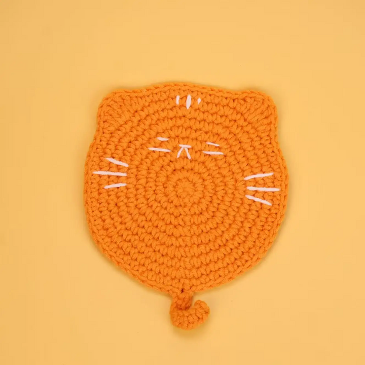 Orange crocheted cat coaster with white stitched whiskers and a curled tail