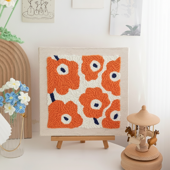 Square orange flower embroidery with blue and white centers displayed on a tabletop easel.
