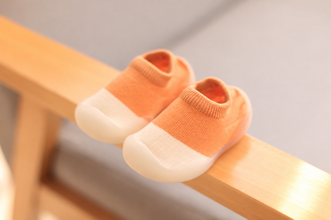 Orange knit baby shoes with cream toe sections resting on a wooden rail.