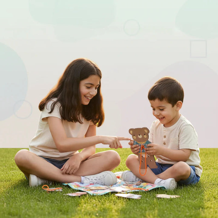 An adult and child sit on the grass while threading a wooden dress-up bear.