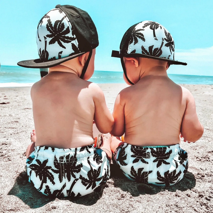 Two children wearing black palm-print caps backward on a sandy beach