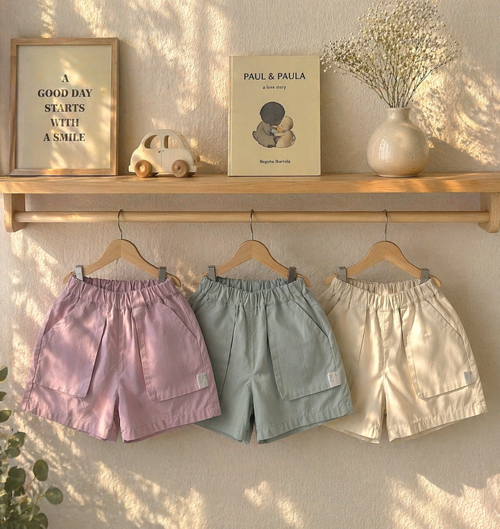 Pink, sage green, and light beige shorts hanging side by side beneath a wooden shelf.
