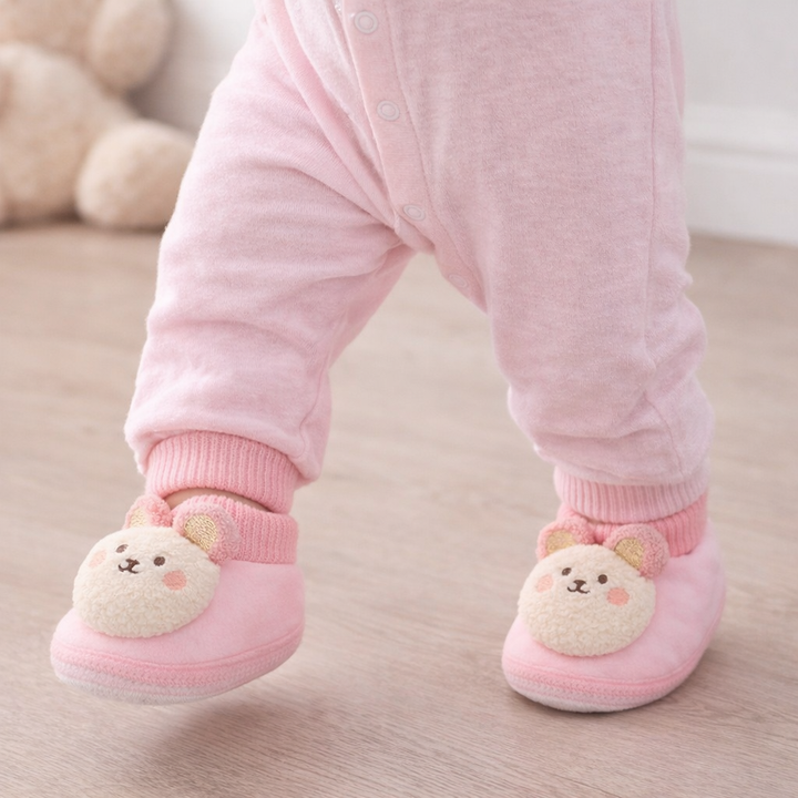 Pink bear baby slippers worn with pink pants on an indoor floor