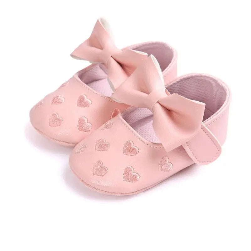 Pink baby shoes with bow straps and heart details, shown at an angle