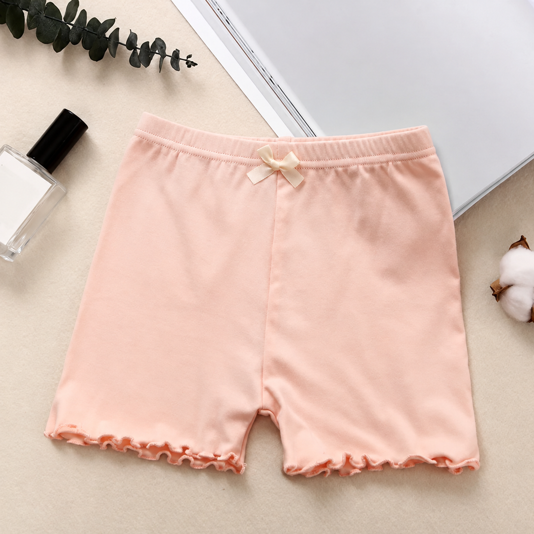 Pink cotton pajama shorts with a cream bow and ruffled hems, laid flat on a tabletop