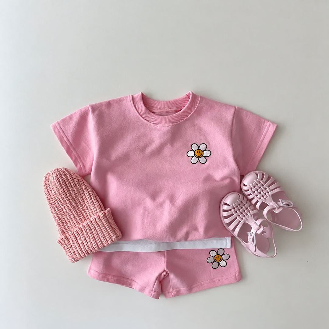 Pink daisy T-shirt and shorts laid out with a pink knit hat and sandals