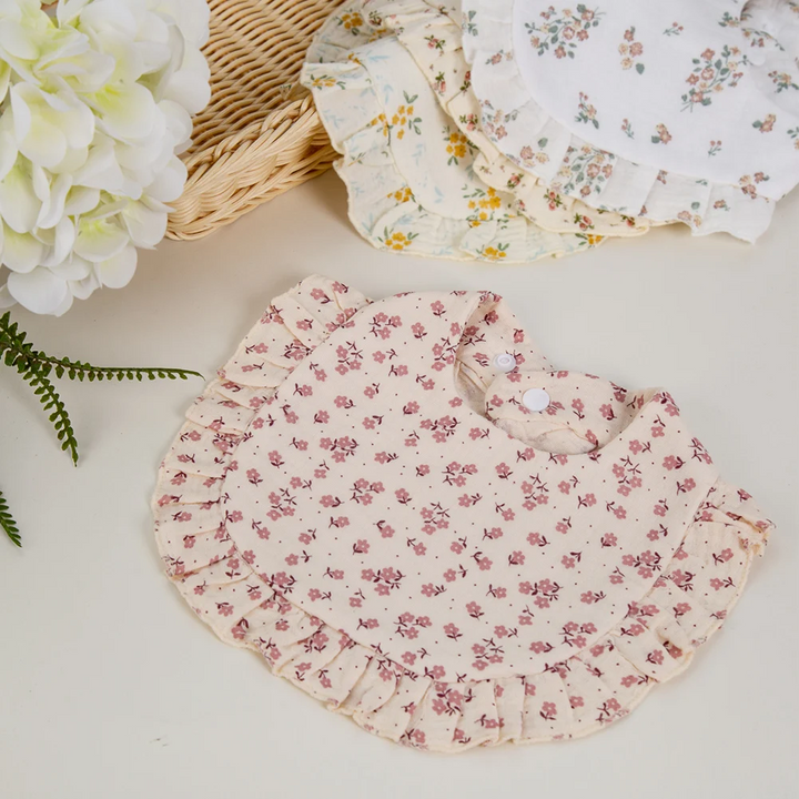 Pink floral ruffled baby bib displayed beside other floral bibs and flowers