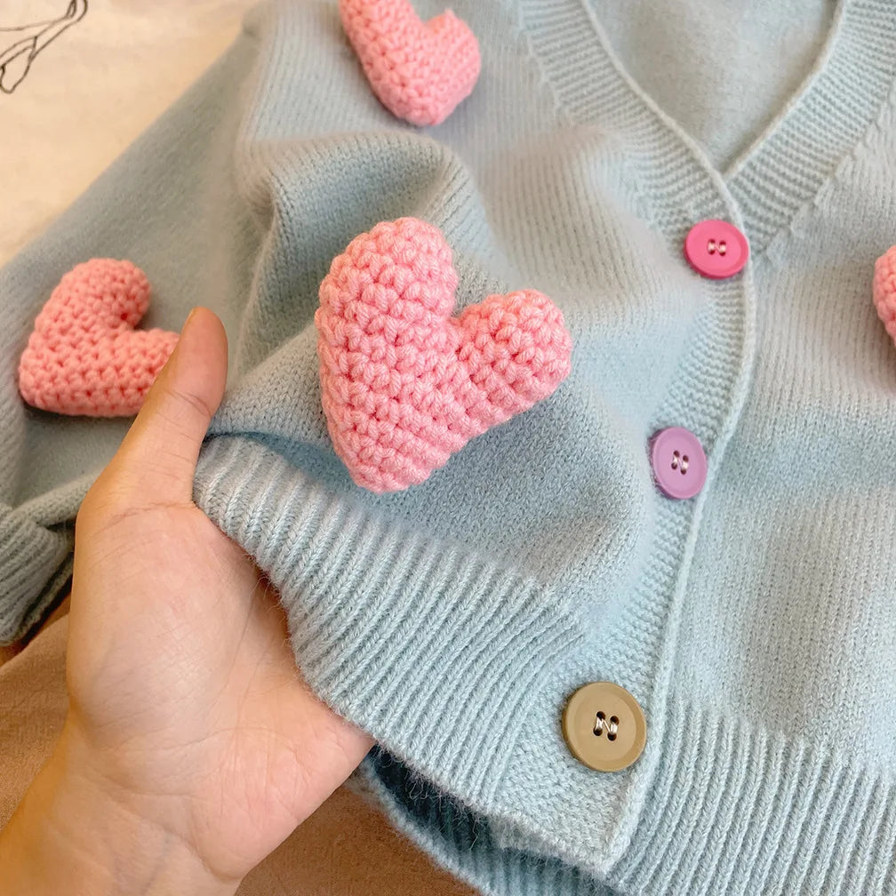 Close-up of a raised pink knit heart, colorful buttons, and ribbed trim on the blue cardigan