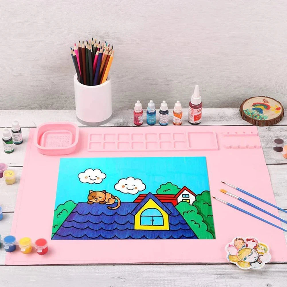 Pink silicone painting mat with a colorful house painting, brushes, and paint supplies on a table.