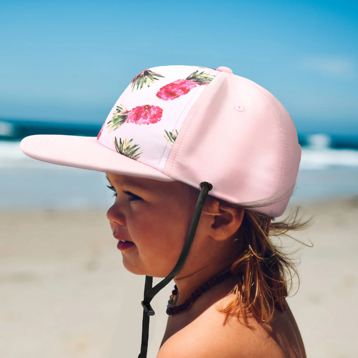 Child wearing a pink pineapple-print cap with a flat brim and black chin strap at the beach