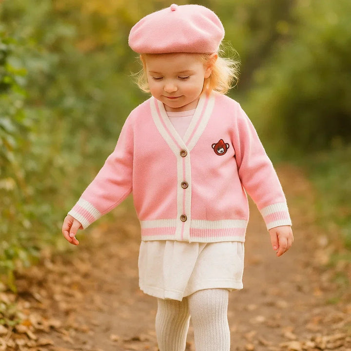 Child wearing a pink teddy bear knit cardigan with cream striped trim and a pink beret outdoors