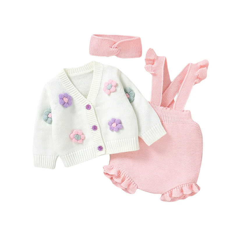 Three-piece pink baby outfit with a white floral cardigan, ruffled overalls and knit headband on white