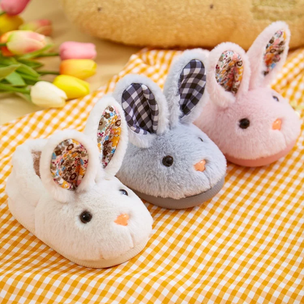 White, gray, and pink plush bunny slippers with upright ears on a yellow gingham cloth