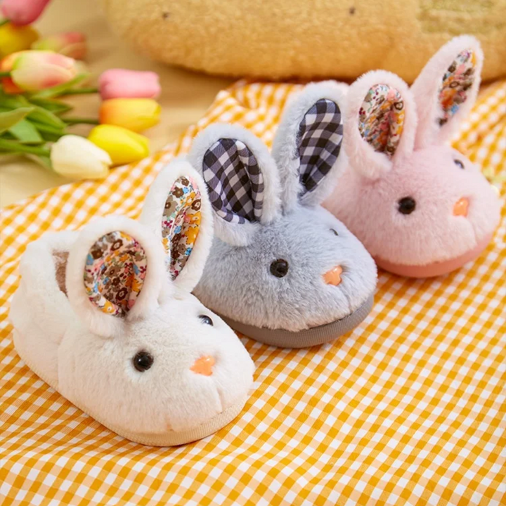 White, gray, and pink plush bunny slippers with upright ears on a yellow gingham cloth