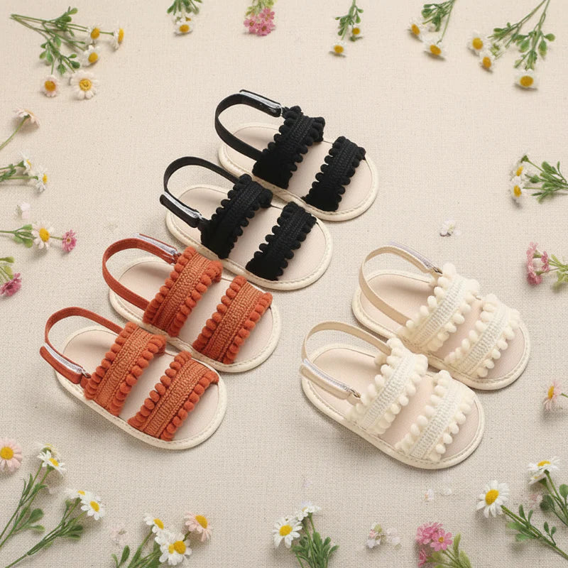 Black, rust-colored and cream baby sandals with pom-pom trim arranged among small flowers.