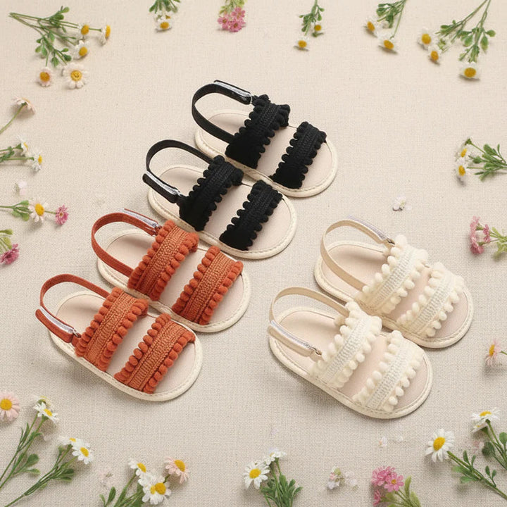 Black, rust-colored and cream baby sandals with pom-pom trim arranged among small flowers.
