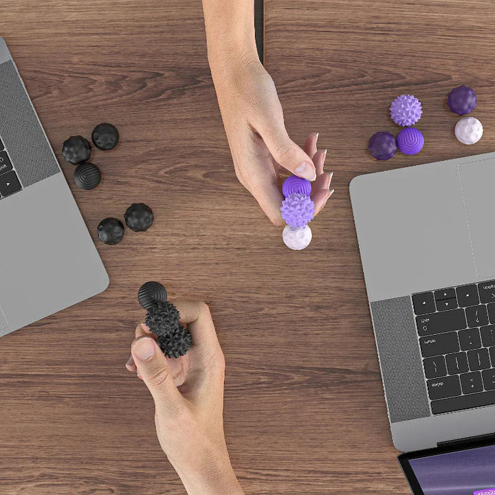 Hands holding purple and black magnetic fidget beads at a desk beside laptops
