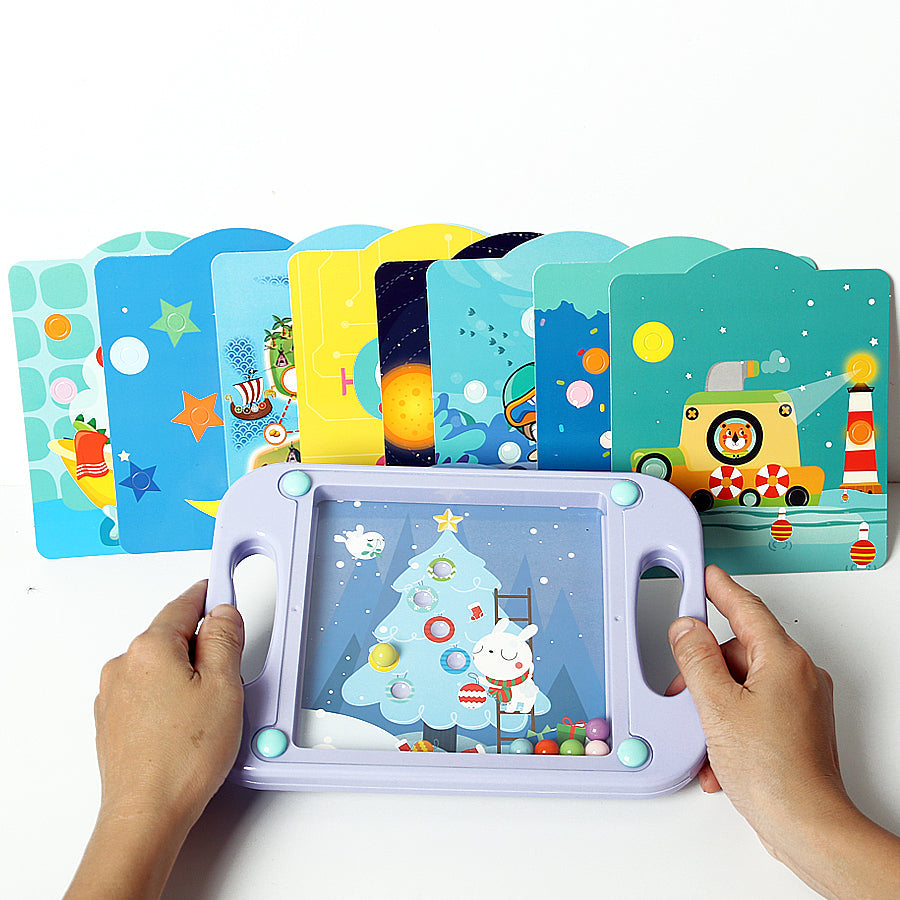 Hands holding a purple ball-maze game with a Christmas tree card and illustrated cards behind it