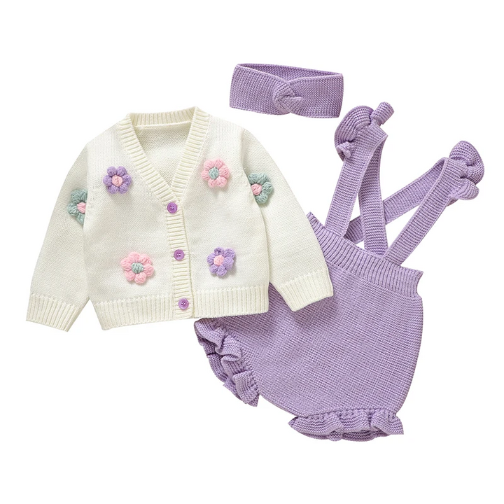 Three-piece purple baby outfit with a white floral cardigan, ruffled overalls and knit headband on white