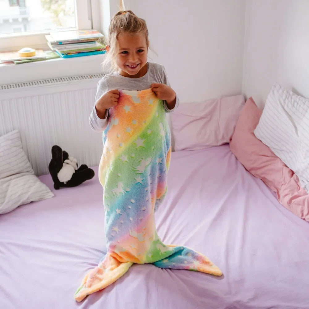Child standing on a bed holding a rainbow mermaid sleeping bag with a tail fin.