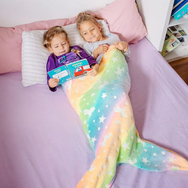 Two children reading on a bed with a rainbow mermaid sleeping bag.