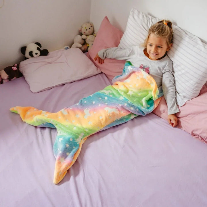 Child relaxing on a bed in a rainbow mermaid sleeping bag with a wide tail fin.