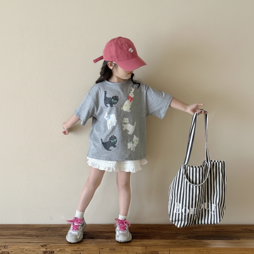 Girl holding her arms out in a roomy gray cat-print tee beside a black-and-white striped tote.