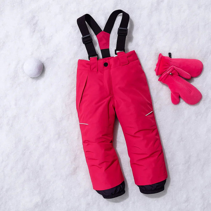 Rosé red ski pants laid on snow with black suspenders, dark lower-leg panels and pink mittens nearby.