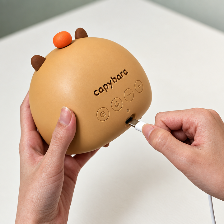 Hands connecting a charging cable to the back of the rounded capybara alarm clock.