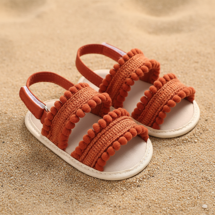 Rust-colored baby sandals with two pom-pom-trimmed front straps and heel straps on sand.