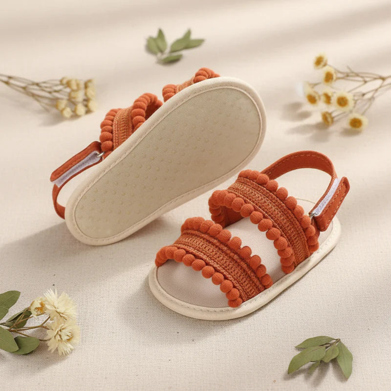 Rust-colored baby sandals with one shoe turned over to show the light-colored sole.