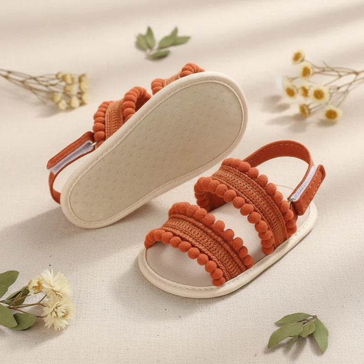 Rust-colored baby sandals with one shoe turned over to show the light-colored sole.
