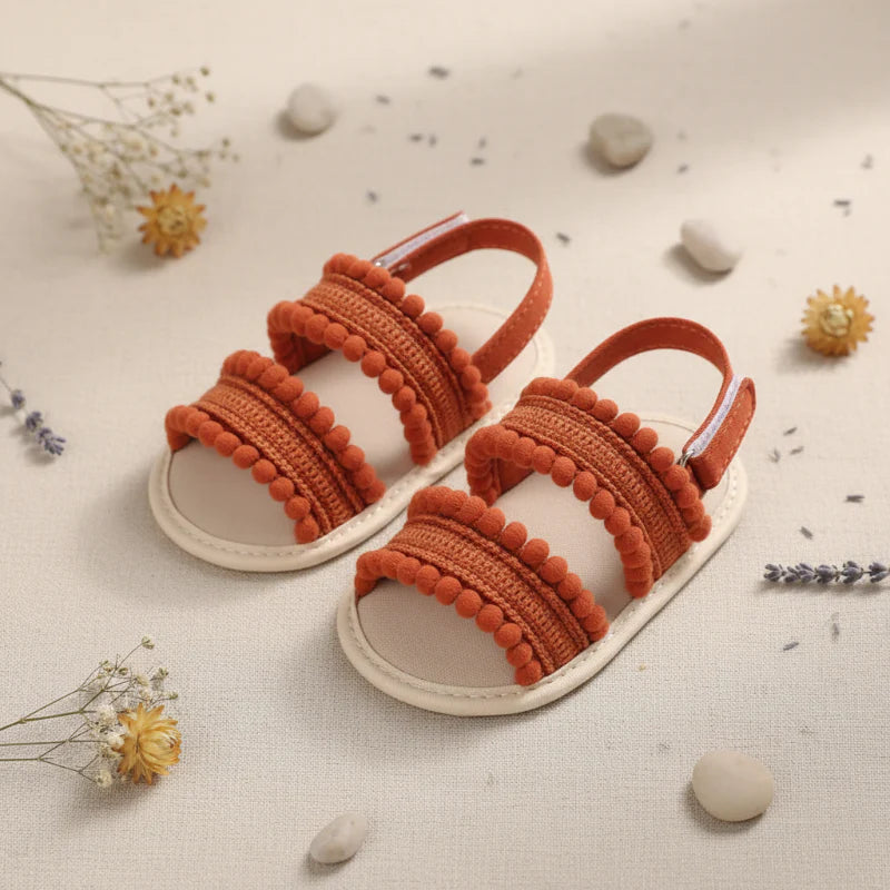Pair of rust-colored baby sandals showing textured front straps and pom-pom edging.