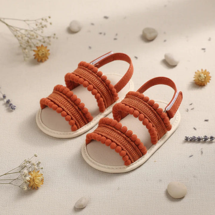Pair of rust-colored baby sandals showing textured front straps and pom-pom edging.